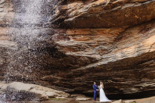 Bride in white wedding dress and groom in blue suit stand under a waterfall