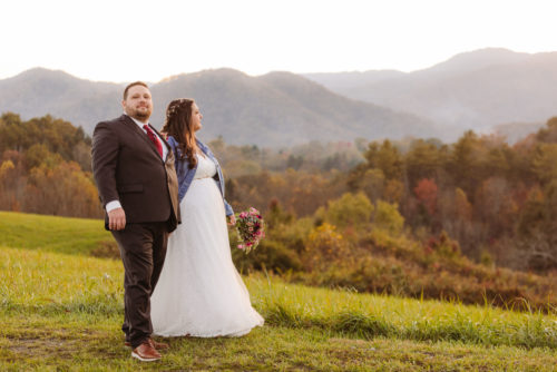 Bride in white wedding dress and jean jacket poses with groom in a suit and red tie in front of blue mountains