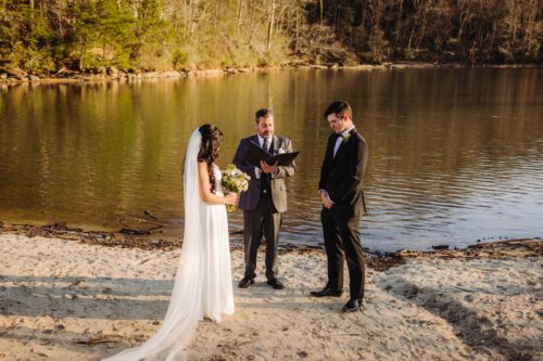Bride in white dress stands with groom in black suit at the edge of a lake as wedding elopement officiant reads 
