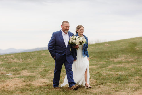 Father escorts Bride in white wedding dress and jean jacket across grassy moun tain knoll 