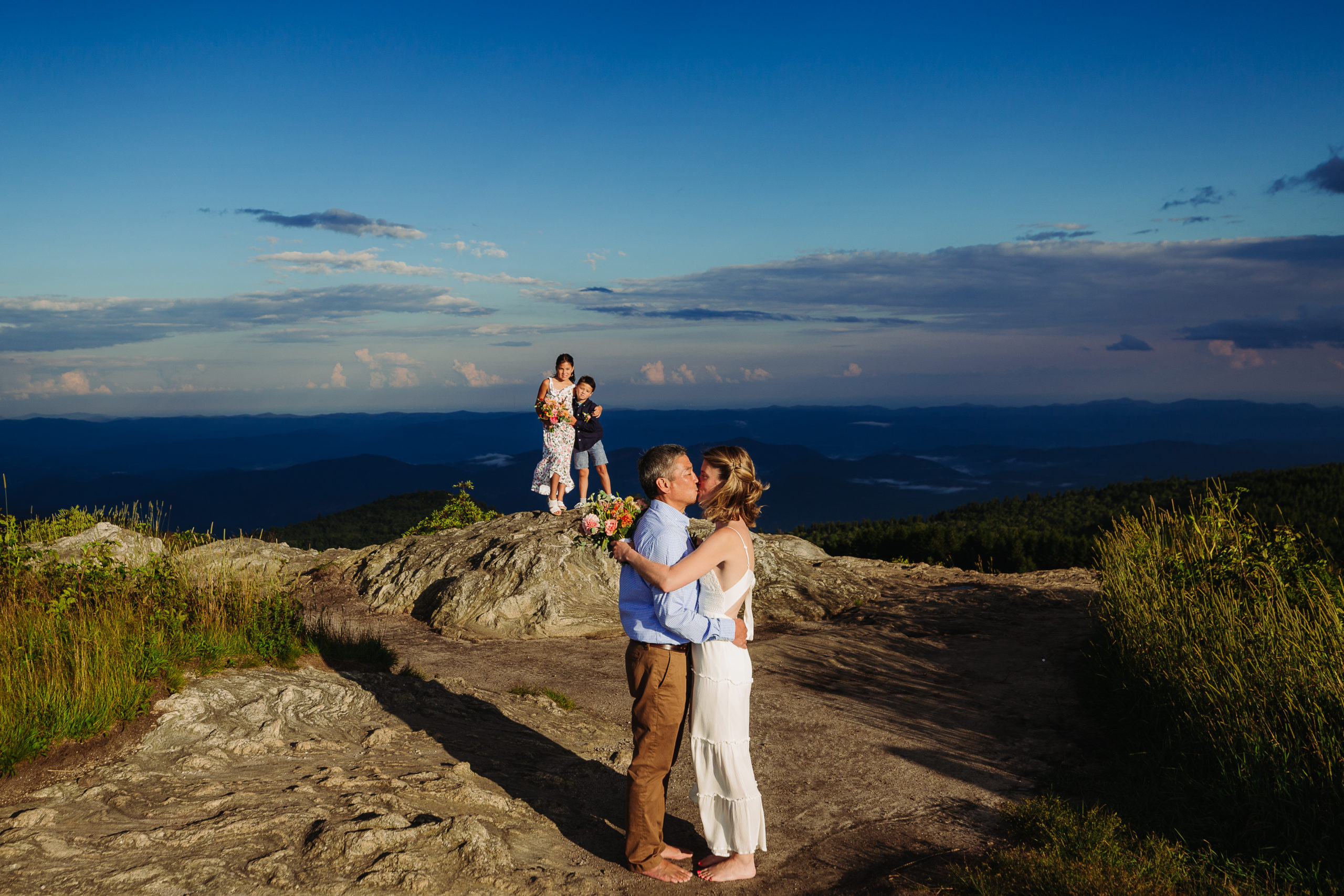 Couple kisses with chilsren behind on rock elopement Elope Outdoors