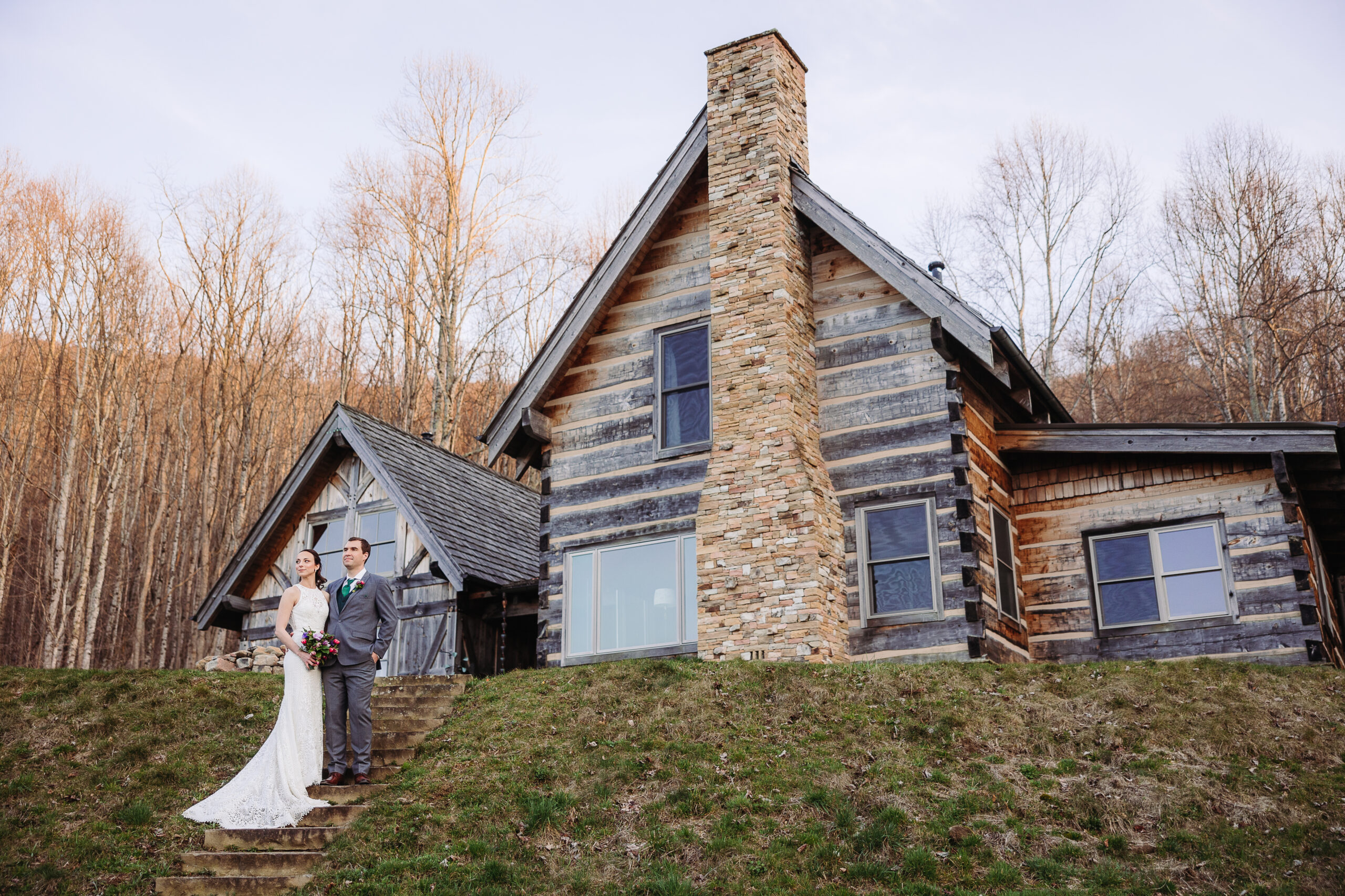 A couple stands on the lawn outside a rustic log cabin (one of Asheville wedding venues) with a stone chimney, surrounded by bare trees in a forested area.