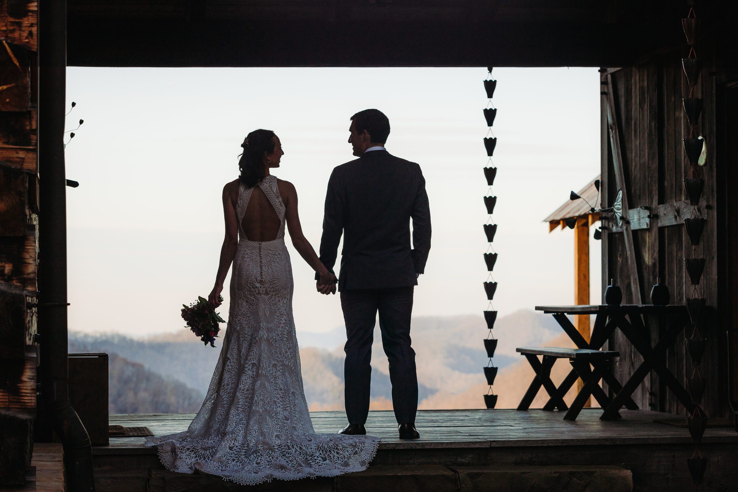 A couple stands hand in hand under a rustic barn structure, looking out at mountain views at an Asheville wedding venues, with the bride holding a bouquet.