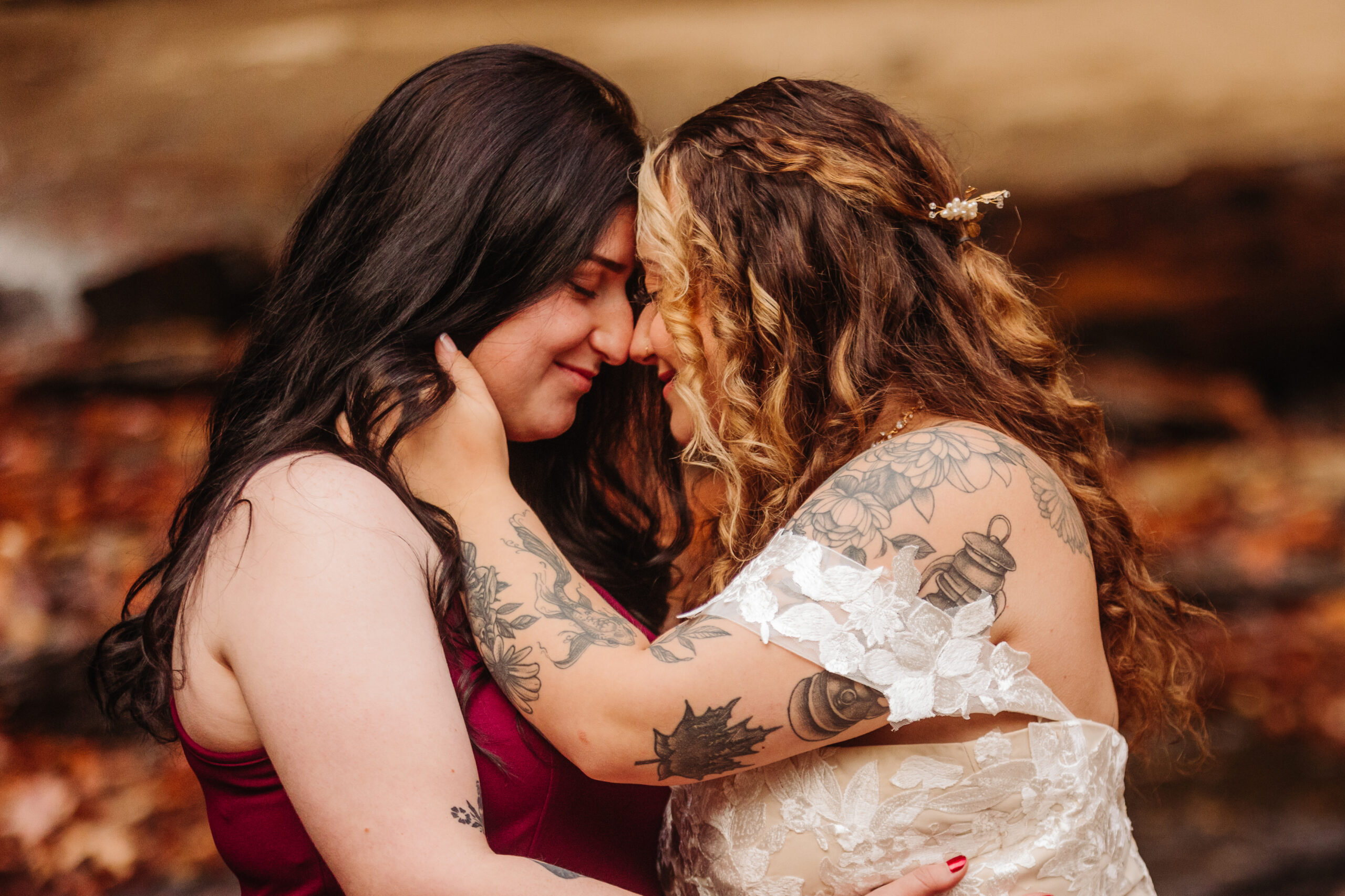 A close-up of two women embracing and touching foreheads, one wearing a white dress and the other a red dress, with an autumnal background.