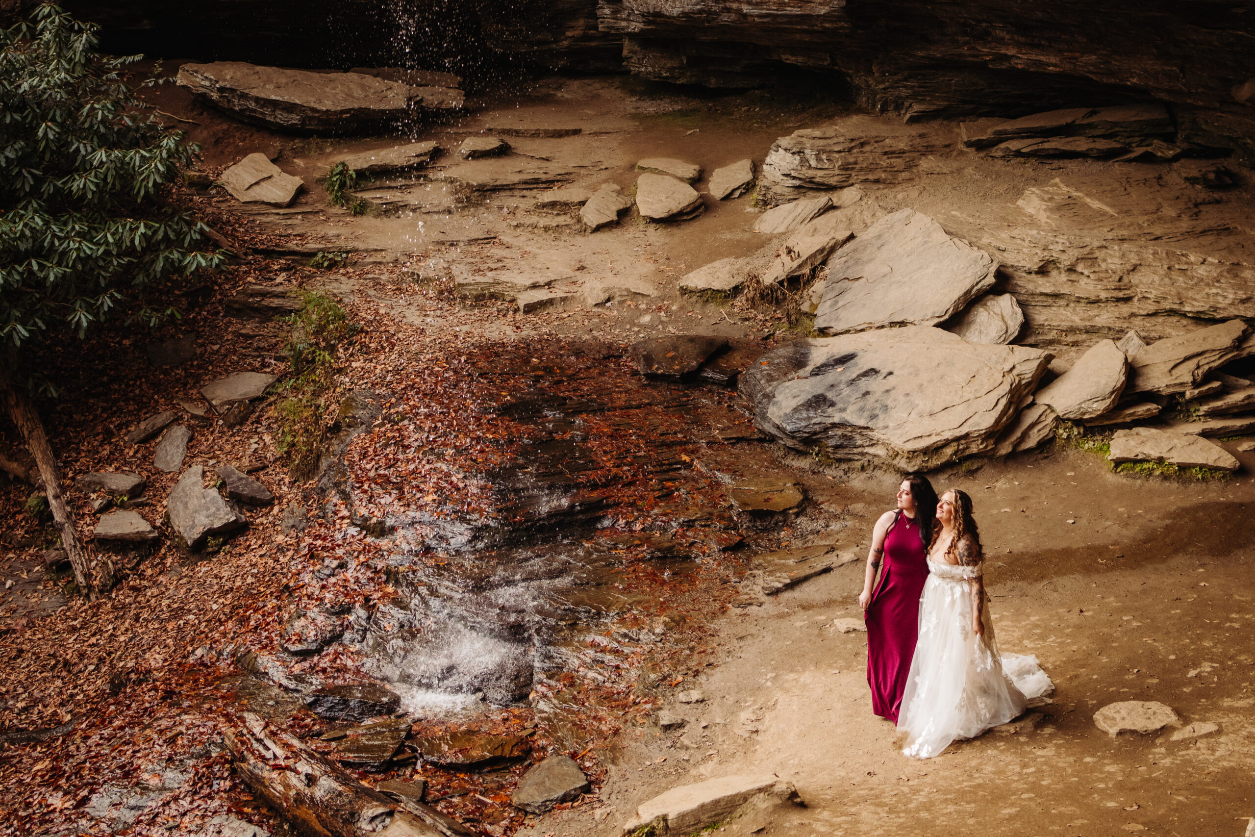 A wide shot of two women standing near a rocky stream bed, one in a red dress and the other in a white dress, surrounded by a natural forest setting.