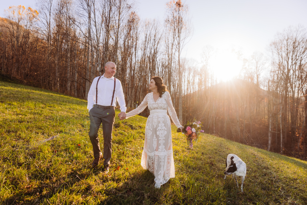 Couple walking hand-in-hand with their dog on a grassy hill during golden hour in Asheville.