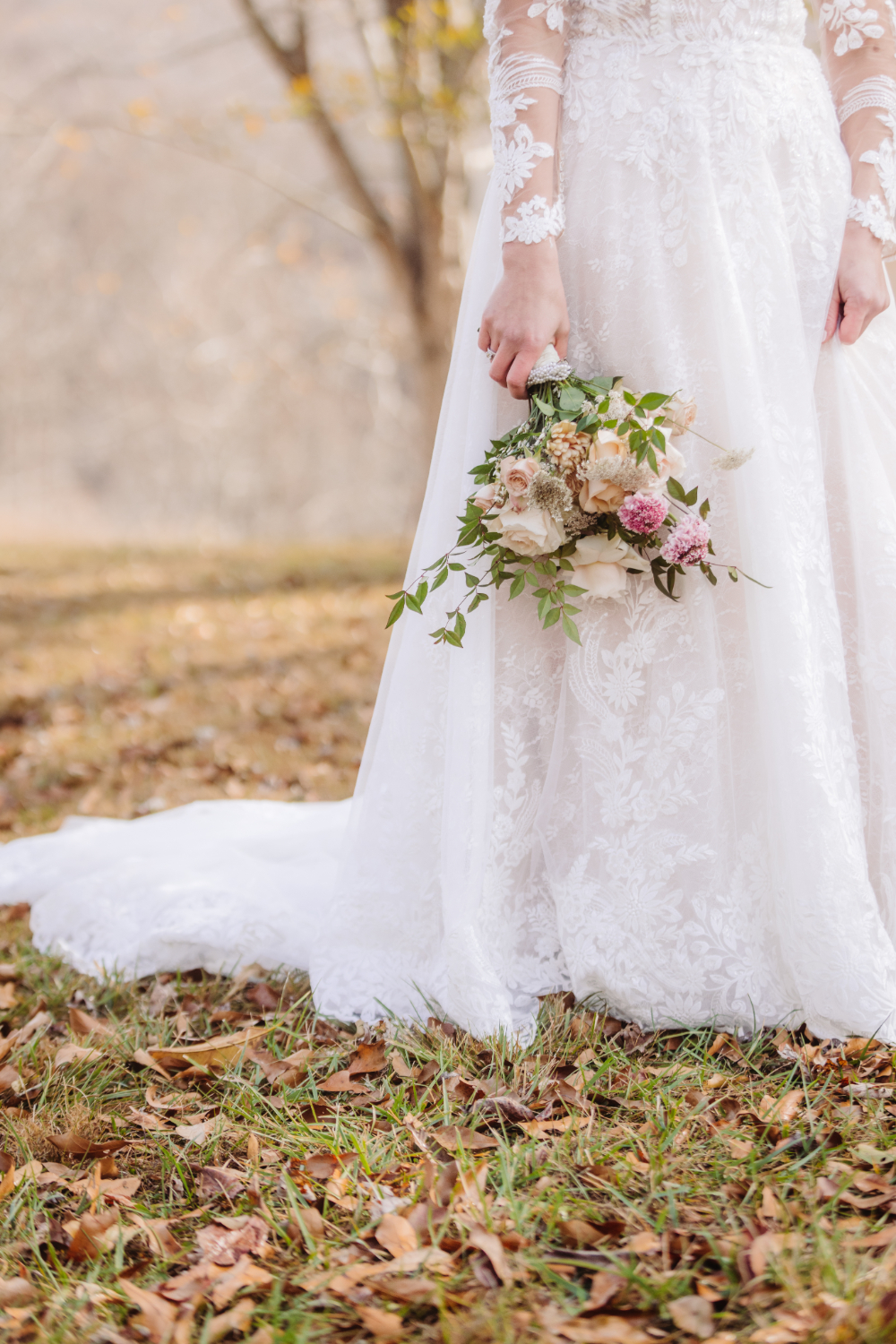 Bride’s lace gown and neutral floral bouquet with greenery in a natural outdoor setting.