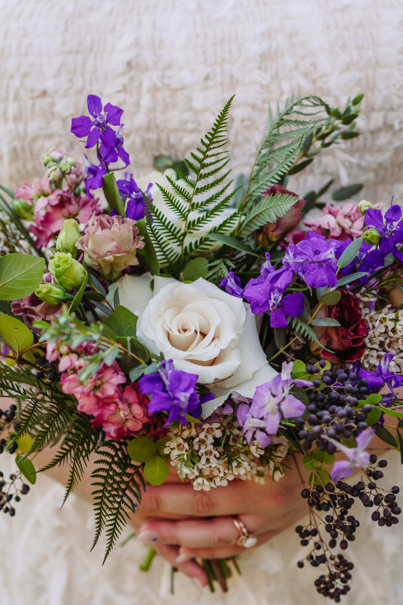 Bridal bouquet featuring purple, pink, and white flowers with greenery.