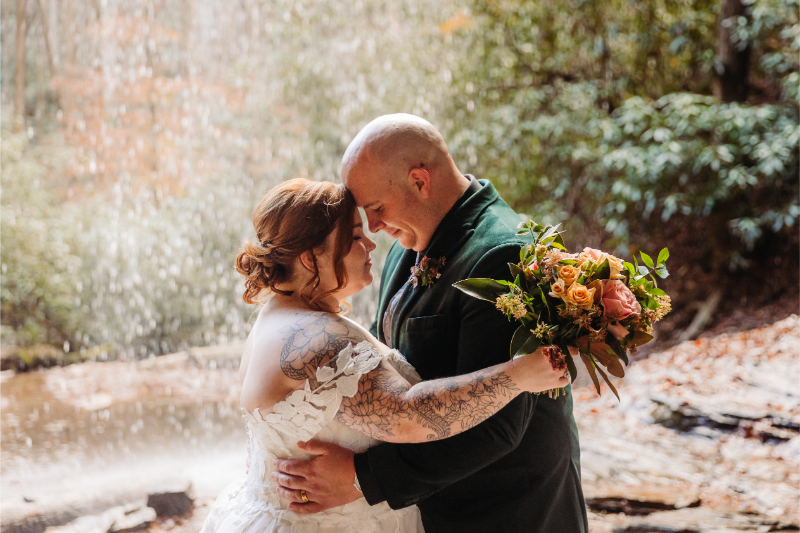 Bride and groom hug in front of waterfall during North Carolina elopement after hiring all-inclusive outdoor elopement planner