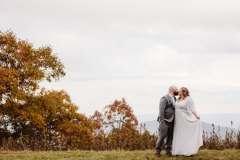 A couple stands together on a grassy overlook with autumn trees and a hazy mountain backdrop under a cloudy sky.