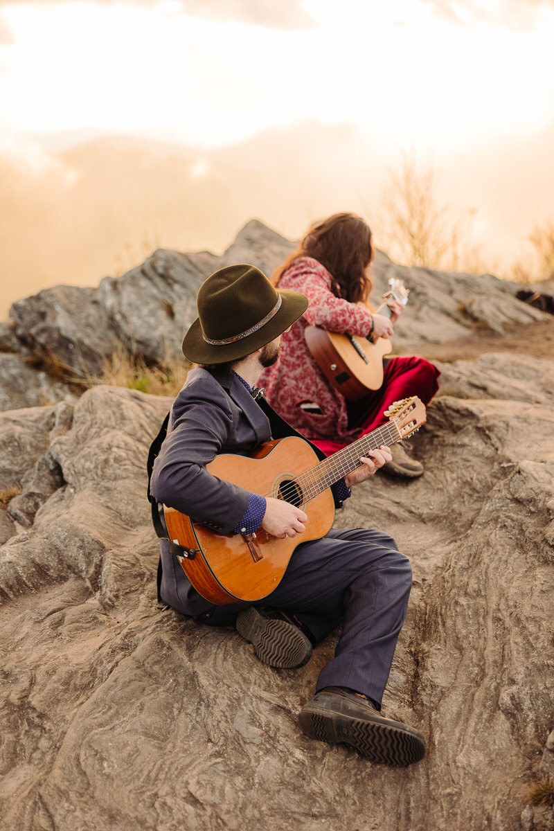 Two bride and groom musicians sit on a rocky overlook, playing guitars with a hazy mountain backdrop and soft light in the distance.