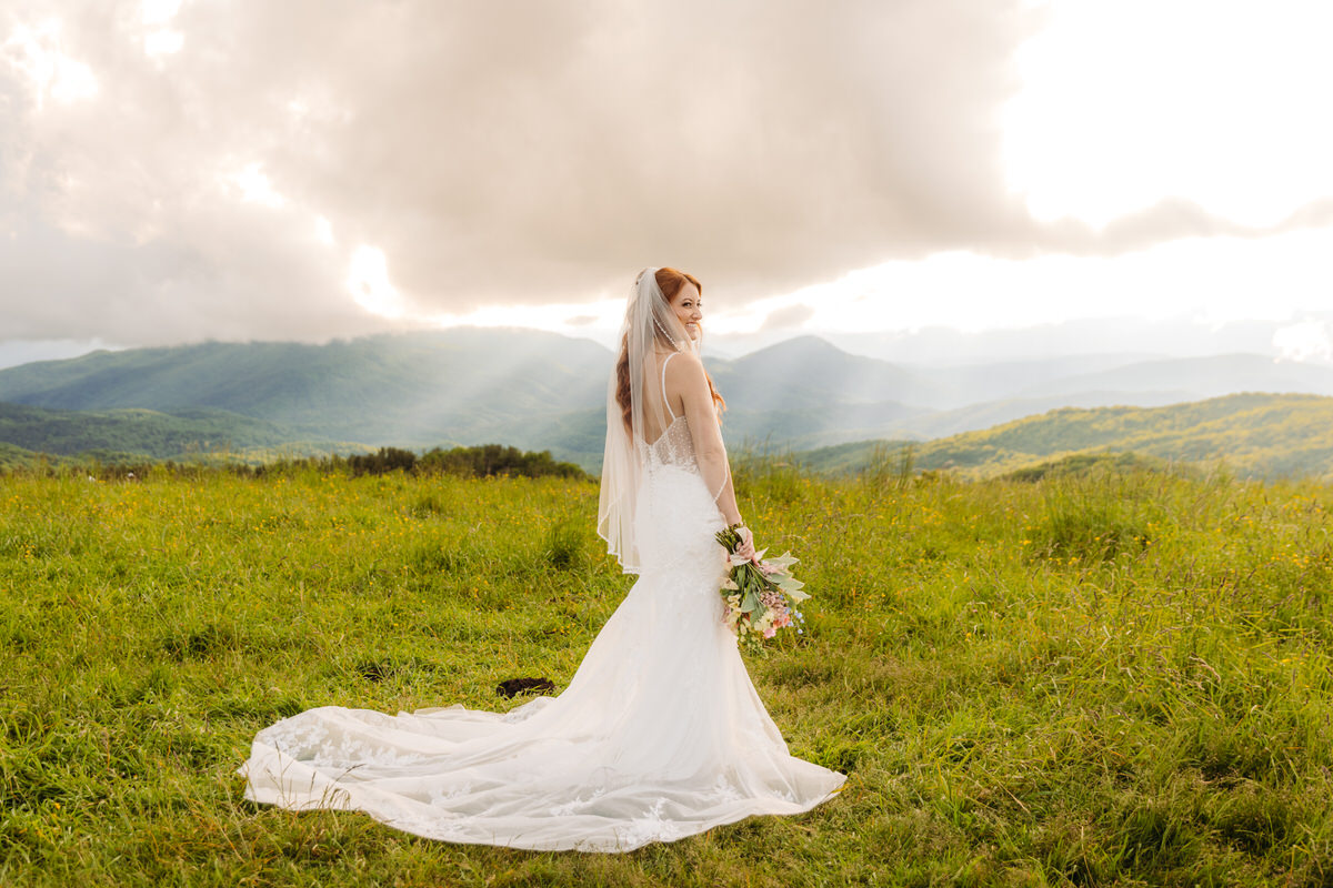 Bride standing on a mountaintop, holding a vibrant summer bouquet with breathtaking mountain views behind her.