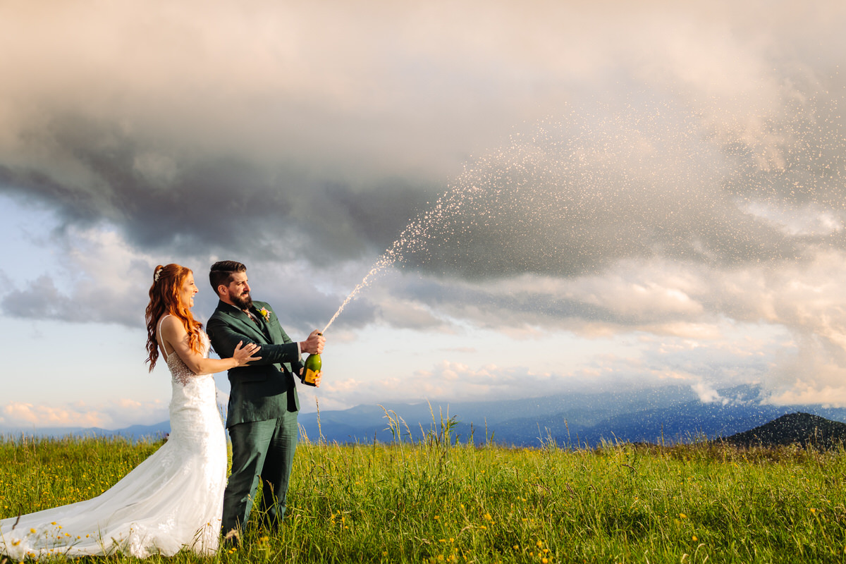 Couple popping champagne on a mountaintop, celebrating their elopement with stunning mountain views behind them.