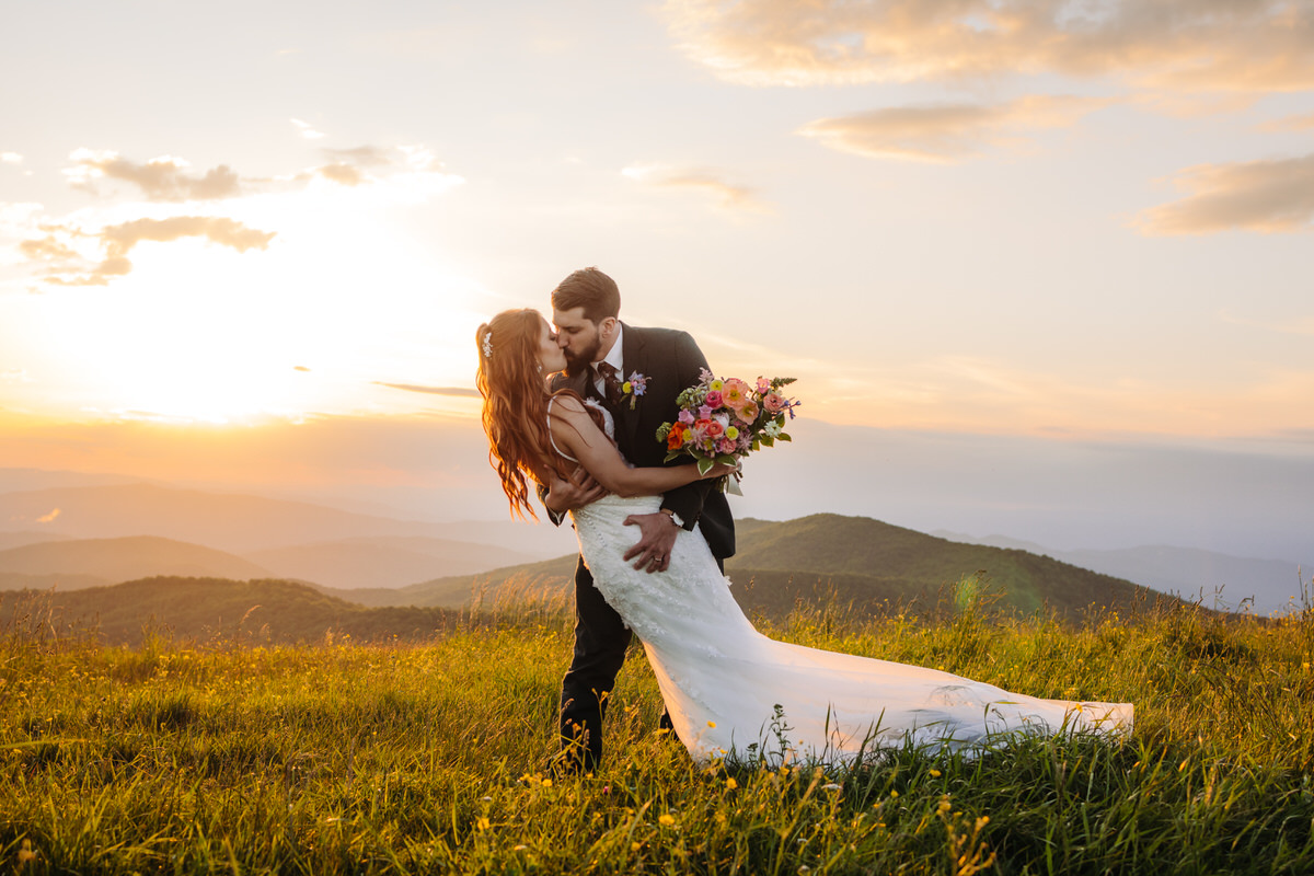 Groom playfully dipping his bride on a mountaintop, surrounded by stunning mountain views.