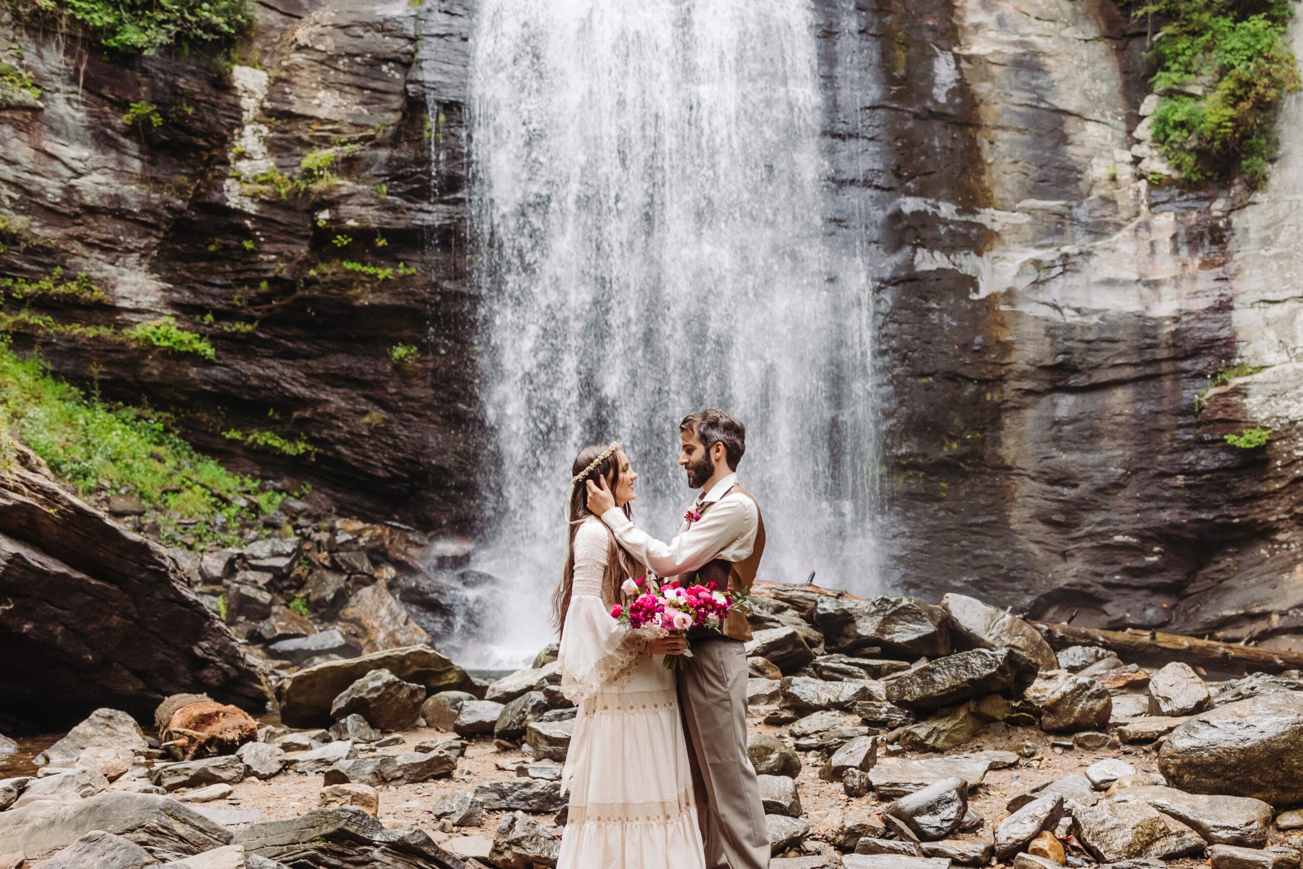 A couple stands barefoot on sun-warmed stones in front of a cascading waterfall, gazing into each other’s eyes. The mist from the falls swirls around them, creating a refreshing and dreamy summer atmosphere.