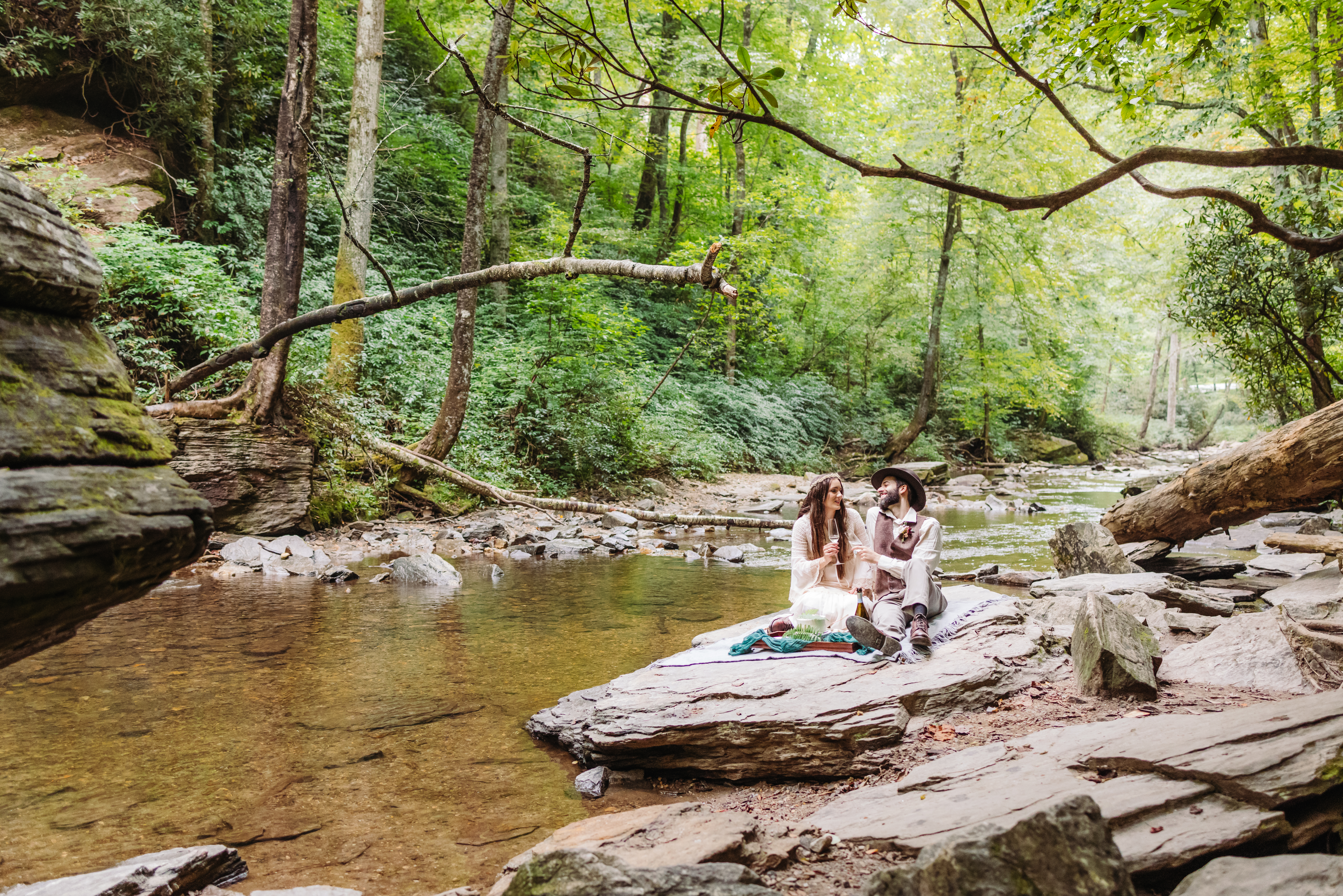 A couple sits on a blanket by the river beneath a waterfall, enjoying a cake and bubbly picnic together, surrounded by lush greenery and the peaceful sounds of nature.