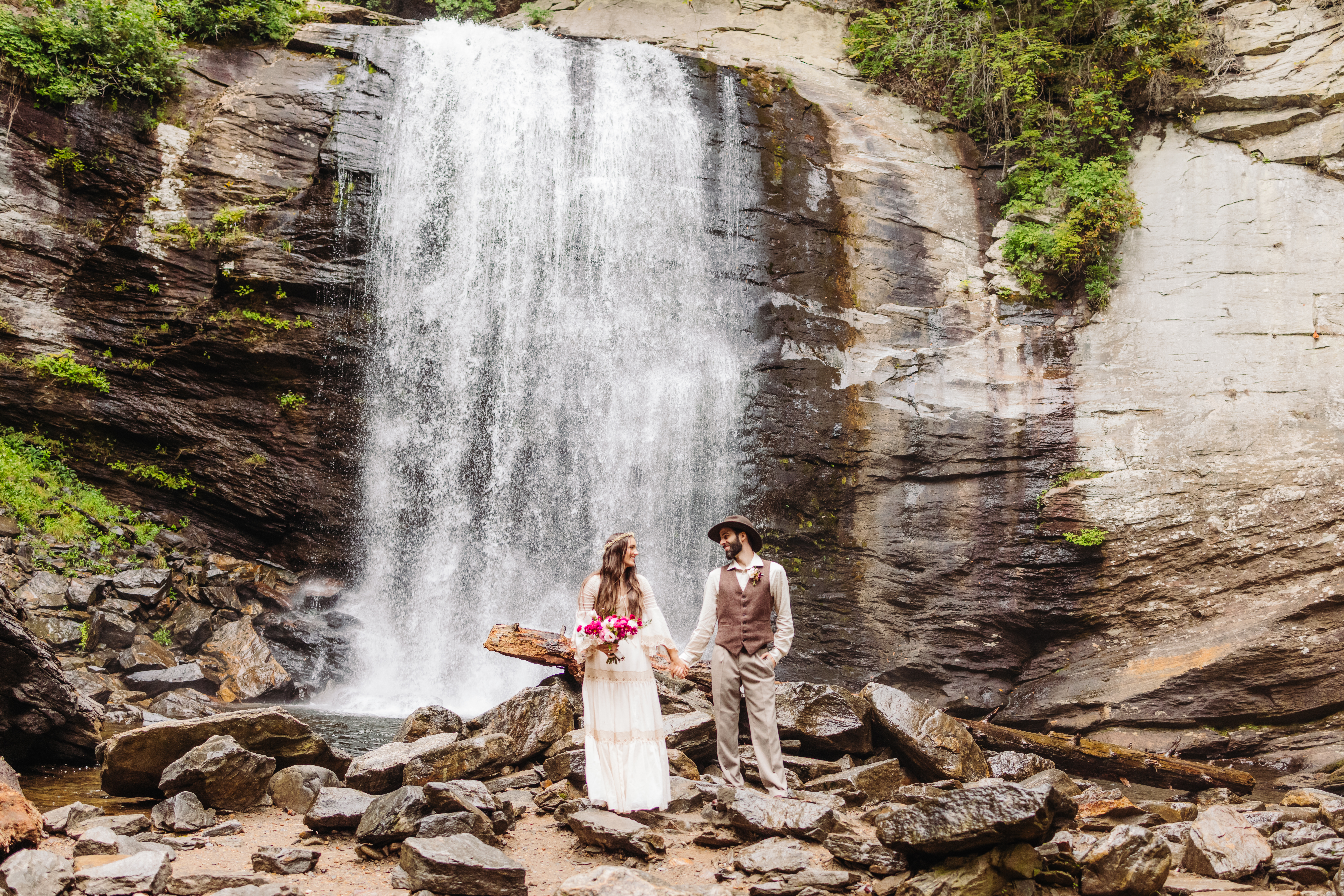 A couple stands hand in hand beneath a cascading waterfall, gazing into each other’s eyes. The lush greenery surrounds them, and the mist from the falls creates a dreamy atmosphere as they share a quiet, intimate moment.