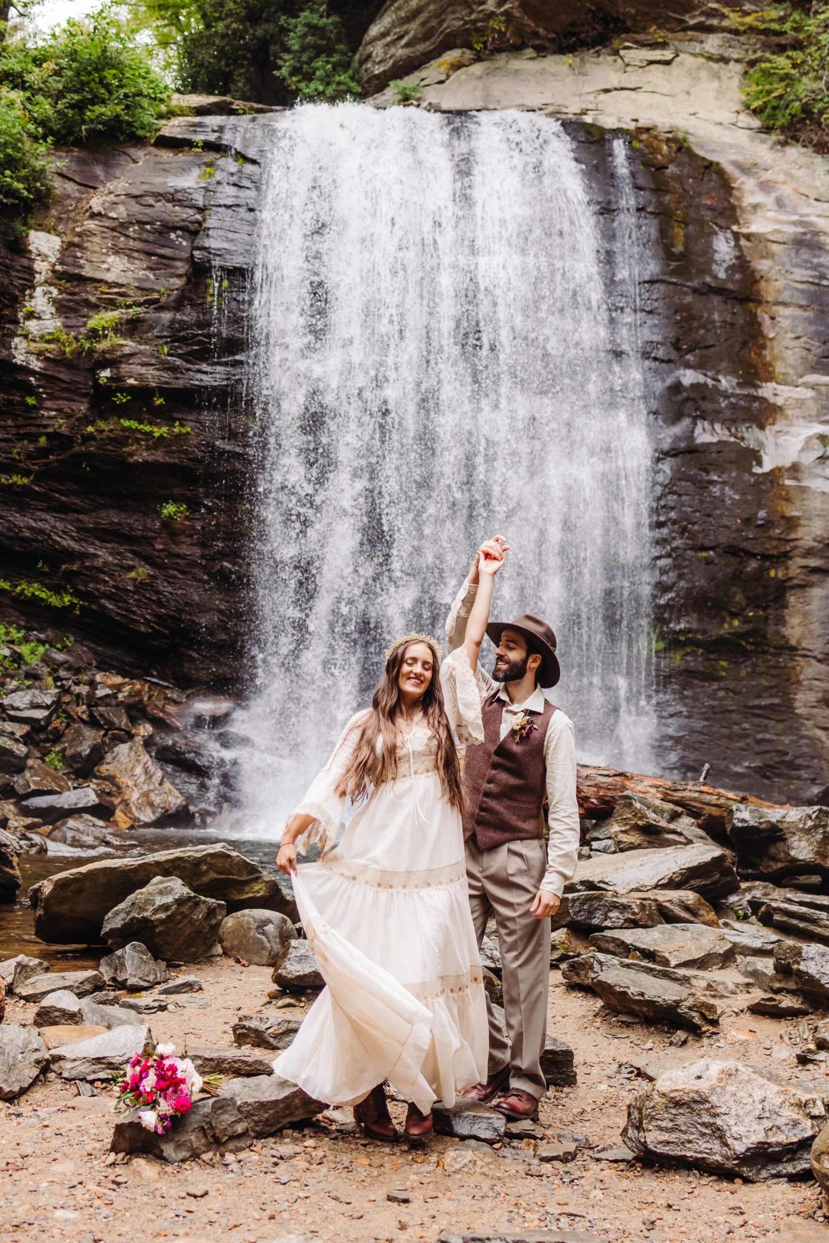 A couple dances in front of a stunning waterfall, with the man giving the woman a playful spin. The mist from the falls surrounds them, creating a romantic and joyful moment in the heart of nature.