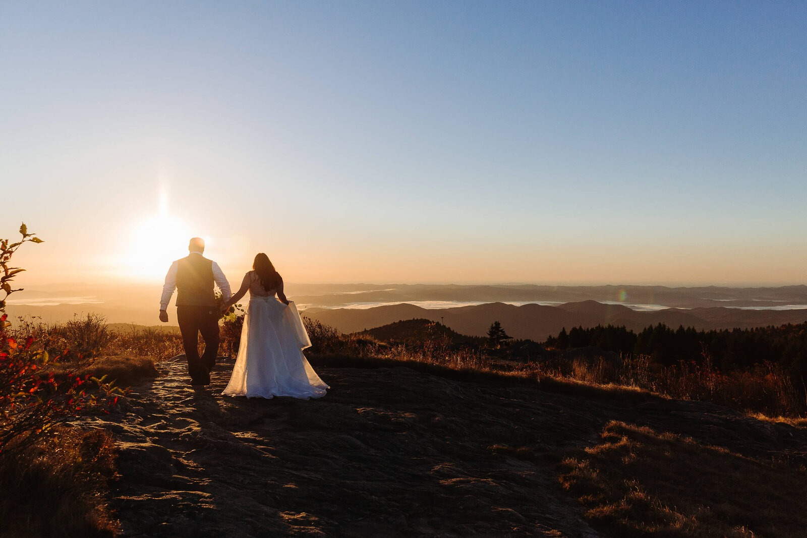 Bride and groom walking hand in hand toward a golden mountain sunset with rolling hills in the distance.