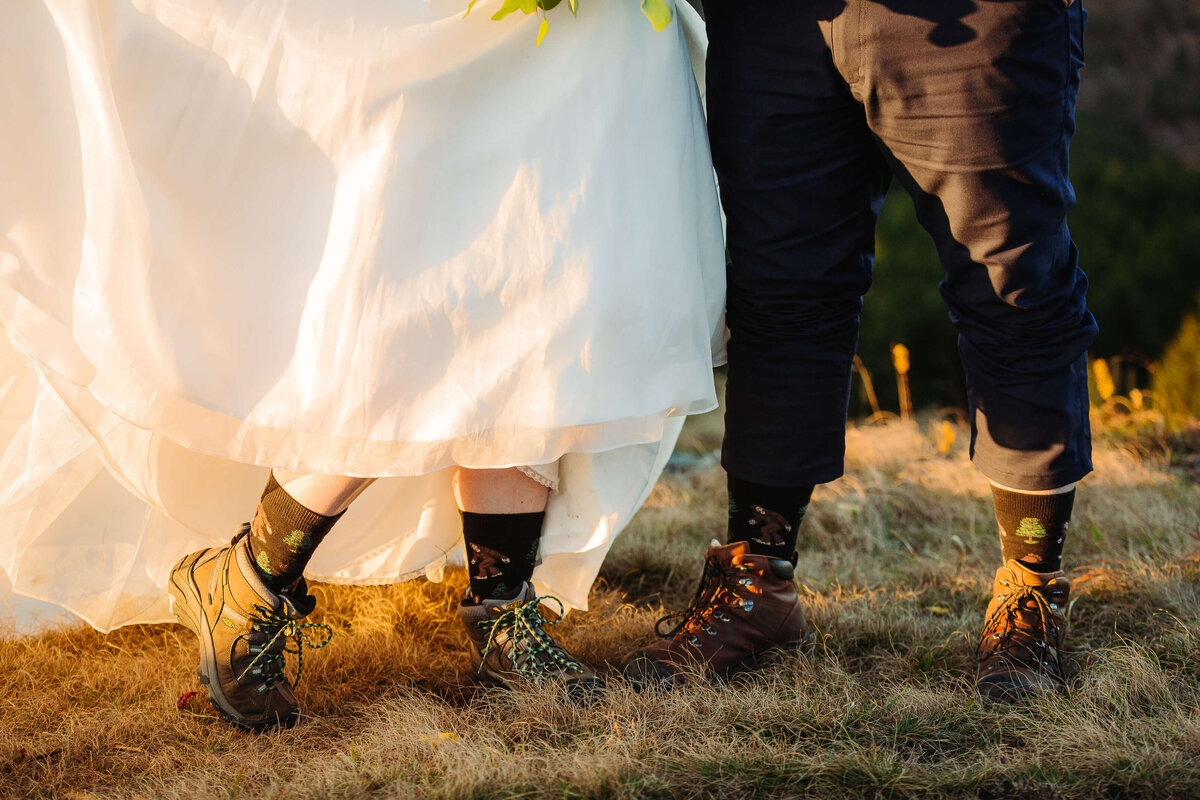 Bride and groom wearing hiking boots and fun socks beneath wedding attire, standing on sunlit mountain grass.