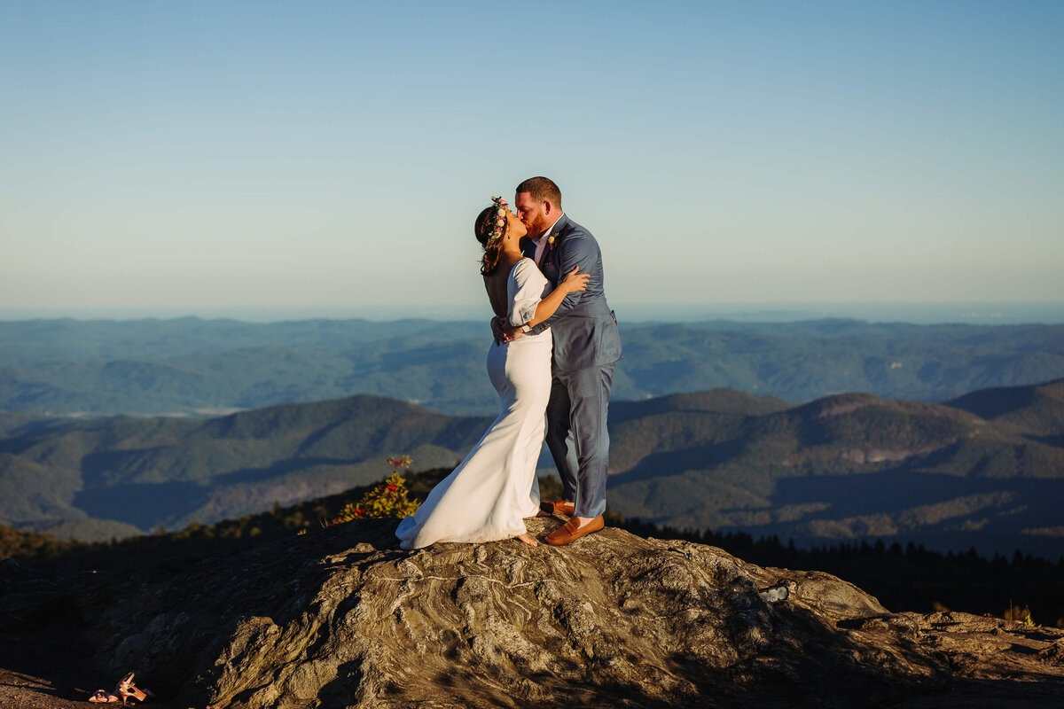 Bride and groom sharing a kiss on a rocky mountaintop with sweeping mountain views in the background.