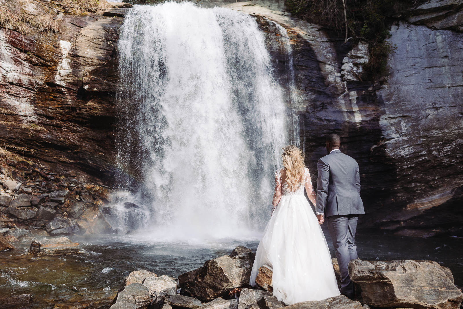 A bride and groom stand hand in hand before a powerful waterfall, showcasing the adventure of an Asheville elopement. Elope in Asheville to experience breathtaking scenery and natural backdrops in every season.