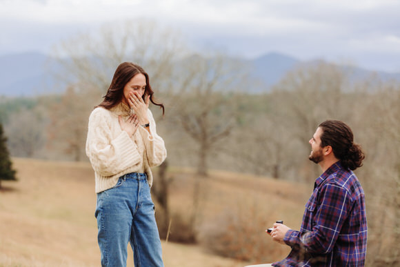 Man proposes outdoors as woman covers her mouth in surprise.
