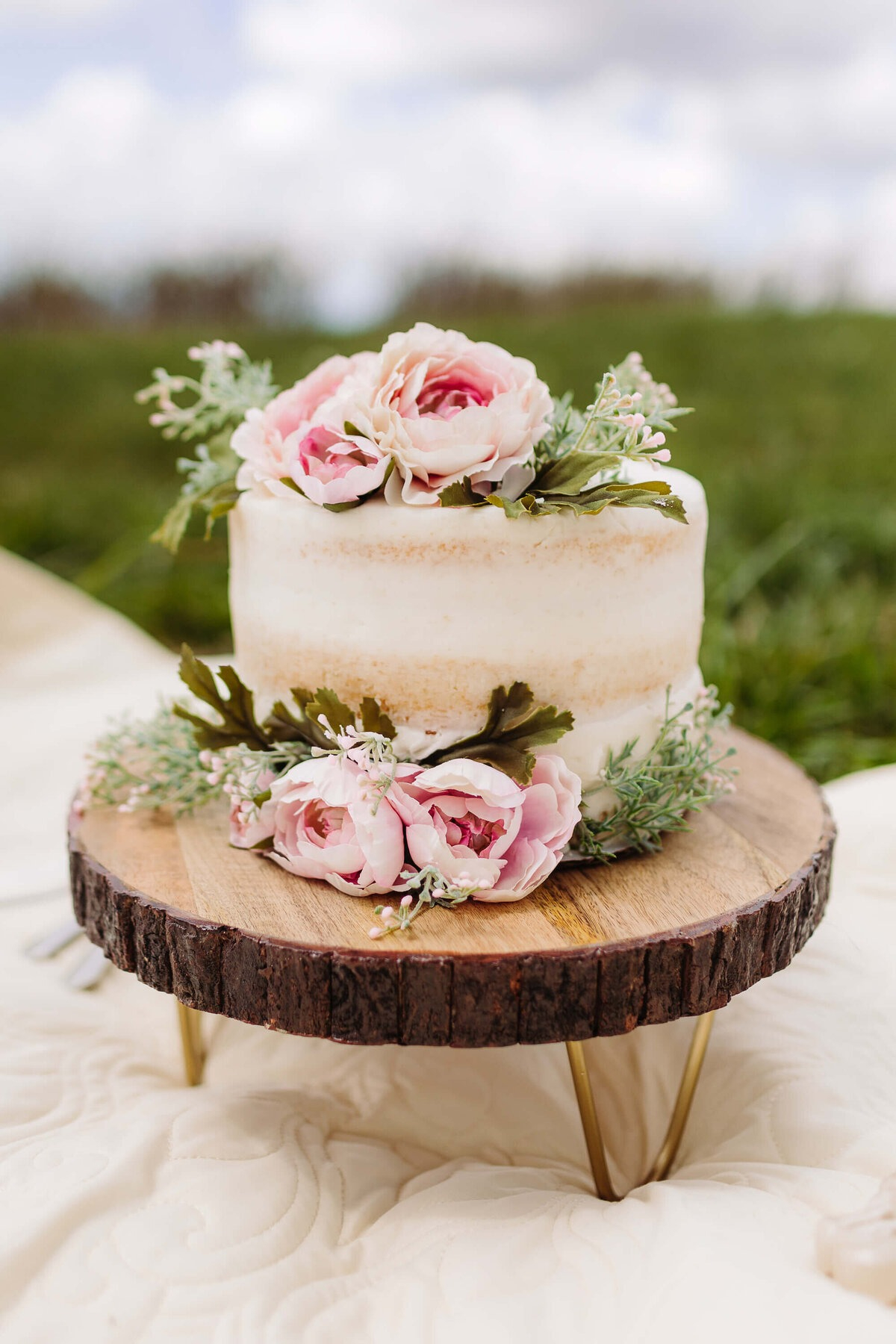 Small semi-naked wedding cake decorated with pink flowers, displayed on rustic wood stand outdoors.