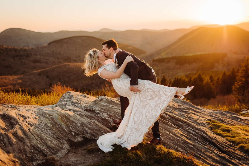 Bride and groom share a romantic sunset kiss on a mountain overlook with golden light all around.