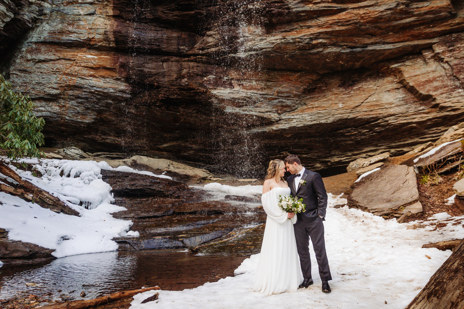 A couple shares an intimate moment under a frozen waterfall during their Asheville elopement. Snow and ice highlight the beauty of winters in Asheville NC.