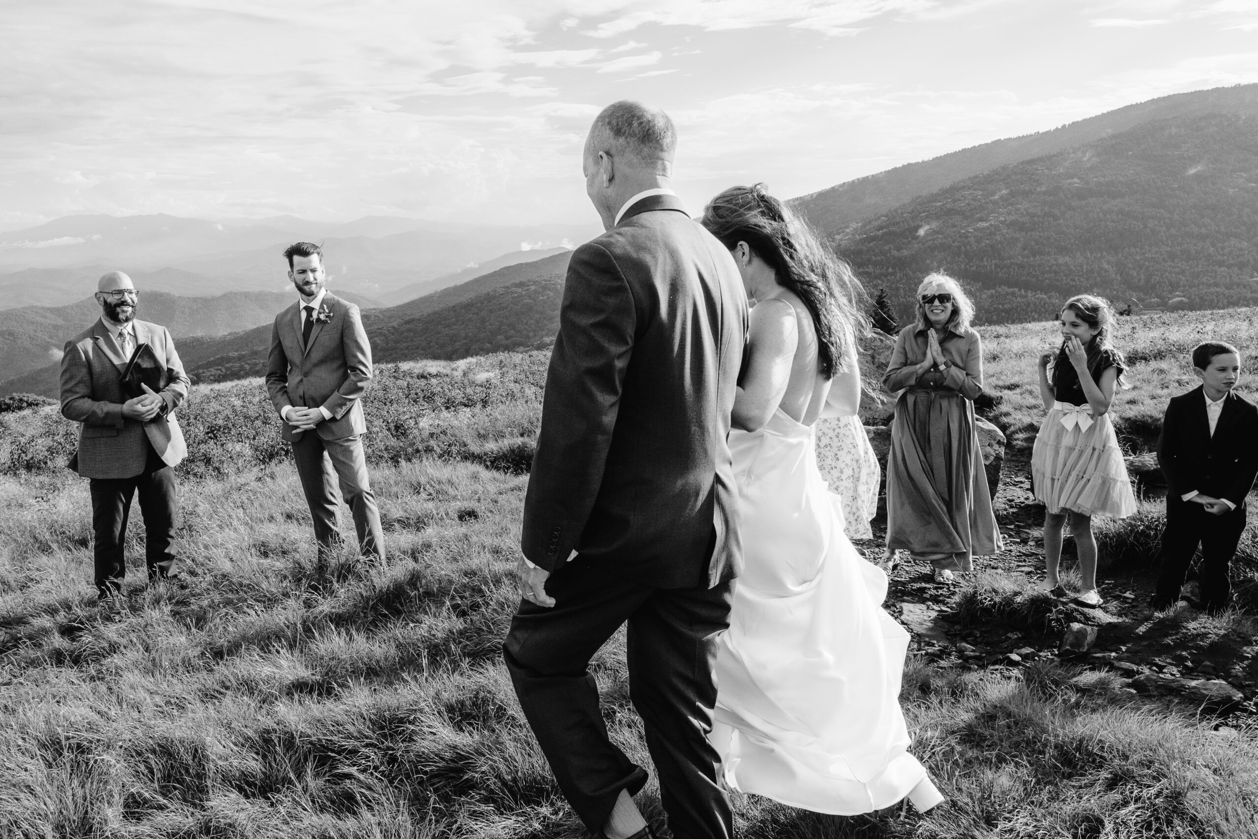 Bride walks with her father toward the groom, officiant, and family during a mountain ceremony.
