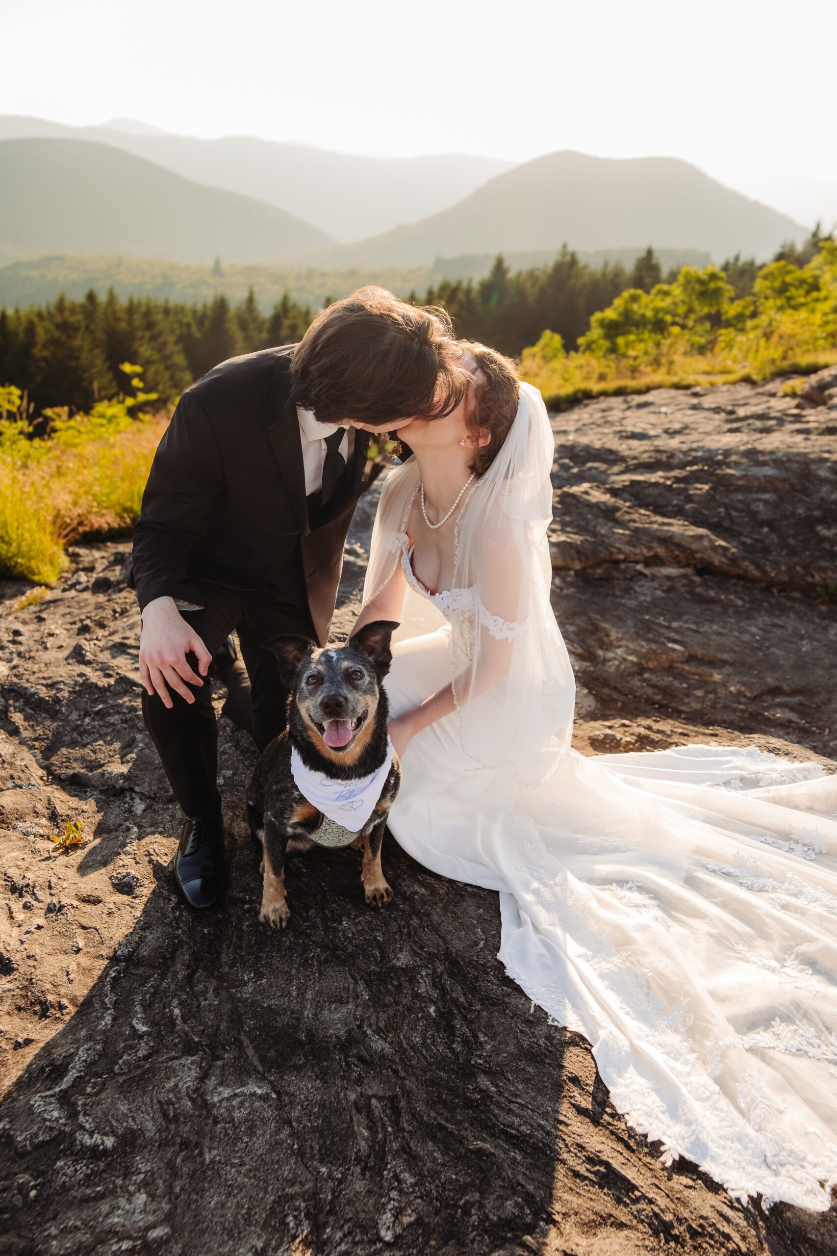 Bride and groom share a kiss on a mountain overlook with their dog wearing a bandana.