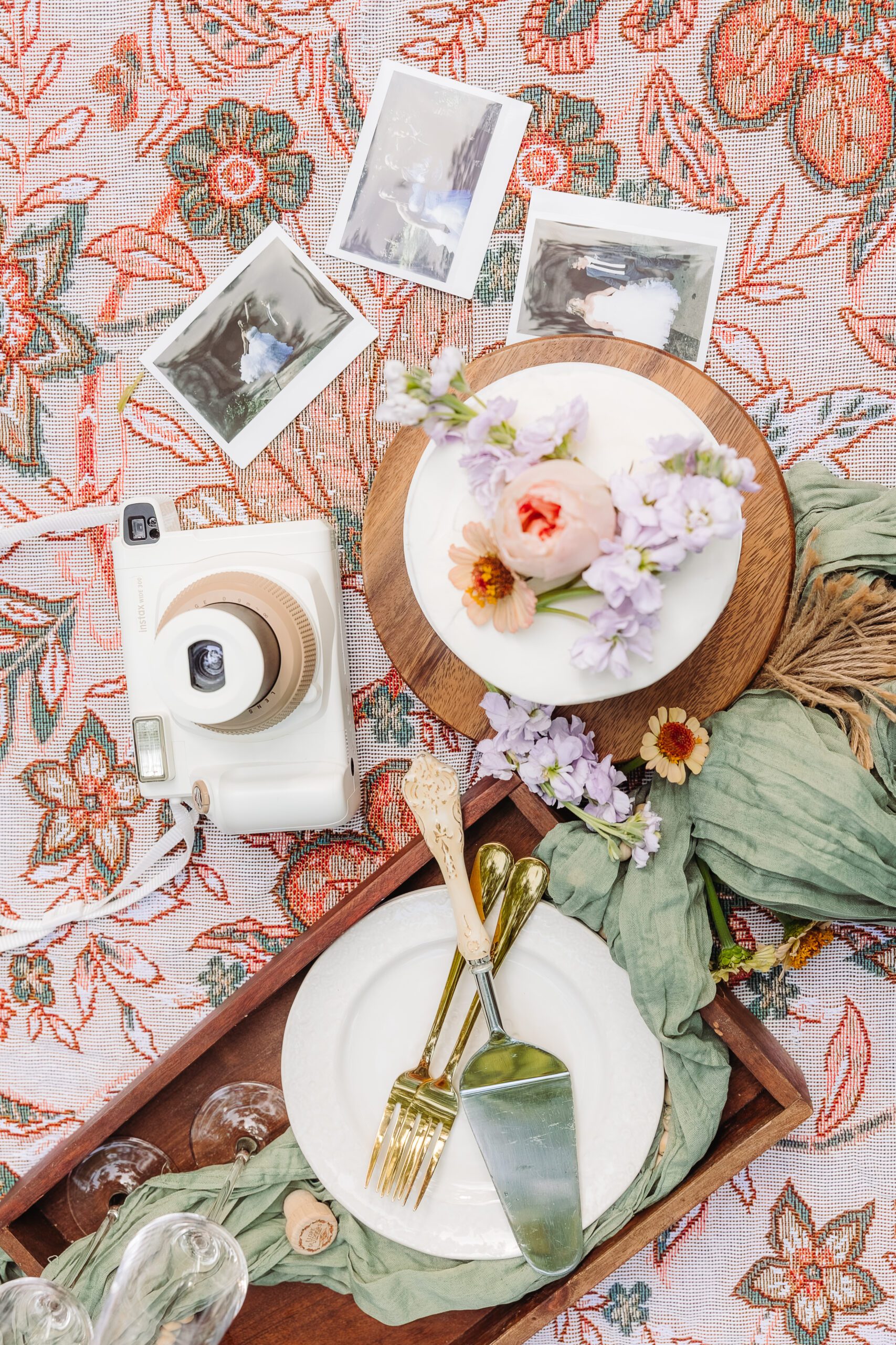 Flatlay of a picnic setup with a floral cake, vintage plates, gold cutlery, camera, and Polaroid photos.