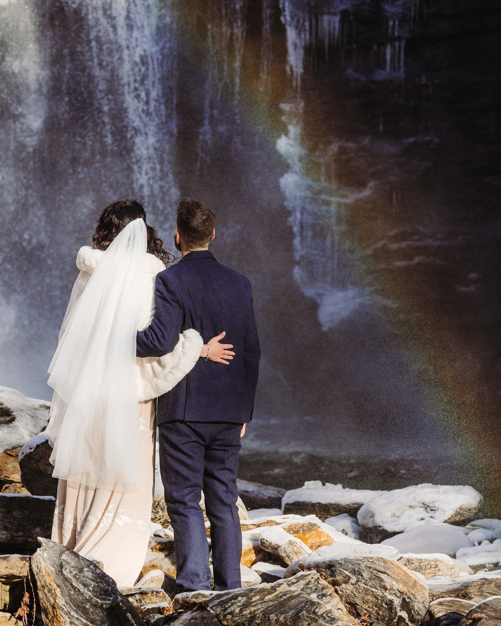 Bride and groom embrace while admiring a rainbow forming in the mist of a winter waterfall.