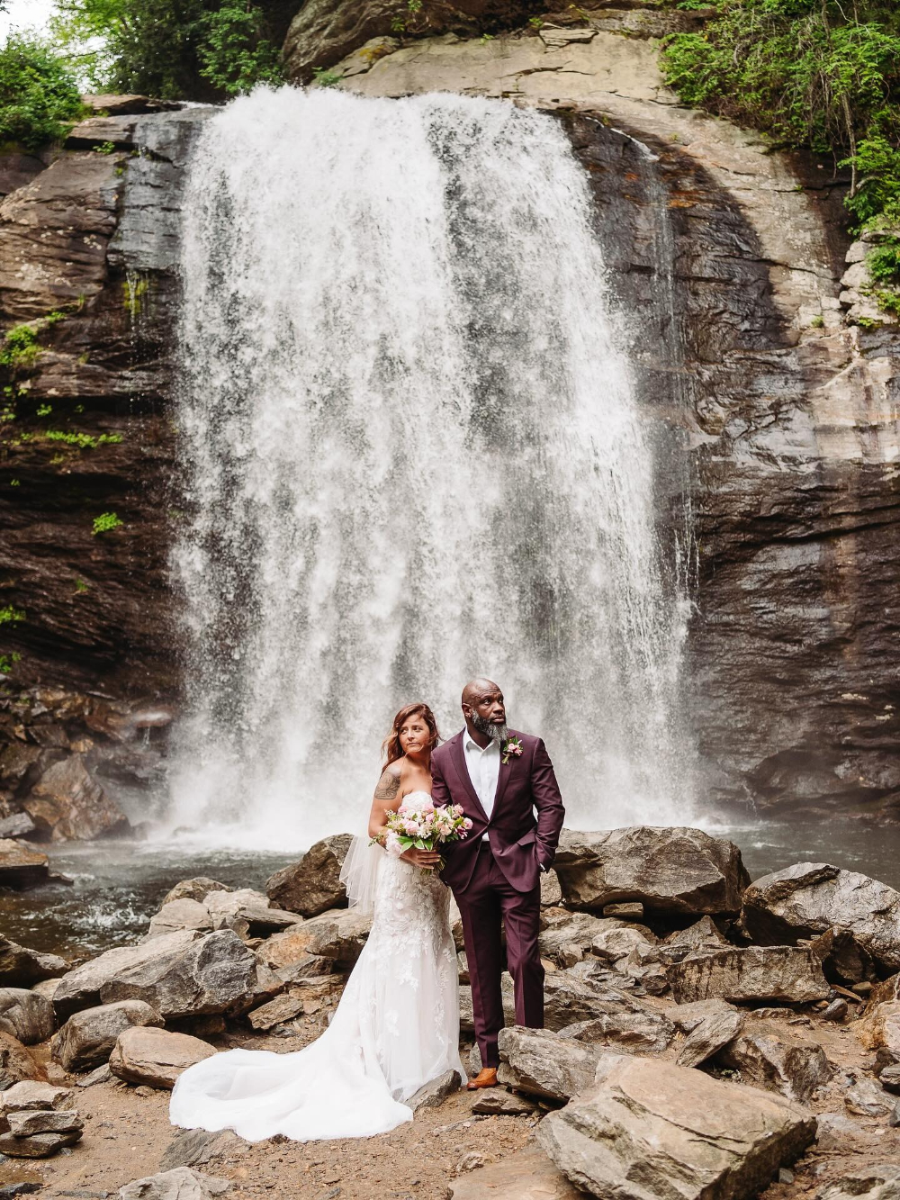 Bride and groom pose on rocks beneath a roaring cascade at one of Asheville’s hidden waterfalls.