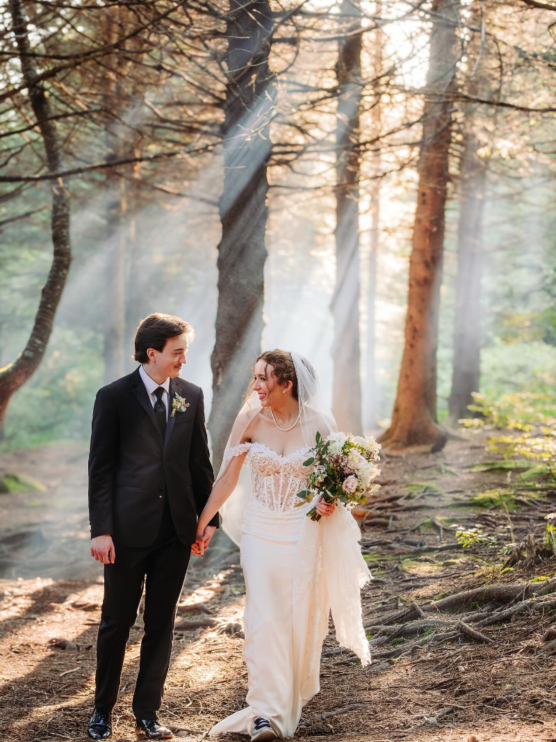 Bride and groom walking hand in hand through sunlit forest, smiling and surrounded by golden morning light.
