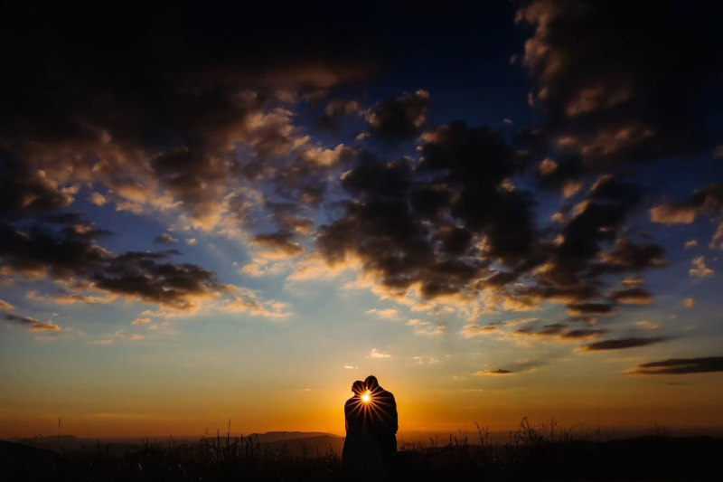 Couple embraces at sunset with the sun shining between them under a dramatic, cloud-filled sky.