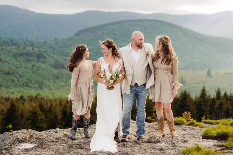 Bride and groom stand with two daughters on a mountain overlook, surrounded by green rolling hills.
