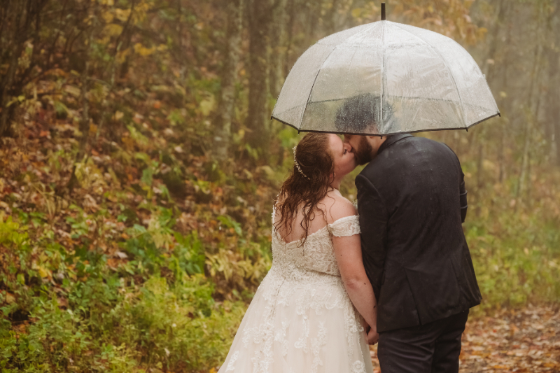 Bride and groom share a kiss under a clear umbrella on a rainy forest path in autumn.