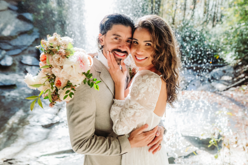Smiling couple poses in front of a waterfall, surrounded by sunlight and flowers—their dream elopement backdrop.
