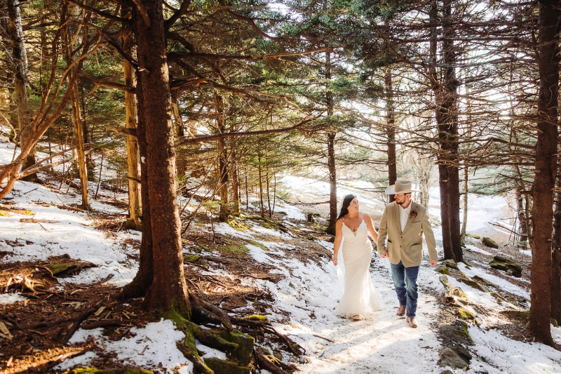 Bride and groom walk hand-in-hand through a snowy forest trail surrounded by tall evergreens and soft light.