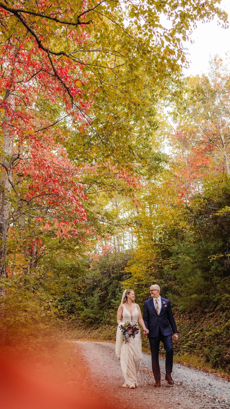 Bride and groom walking down a forest path lined with vibrant fall foliage in warm, golden light.