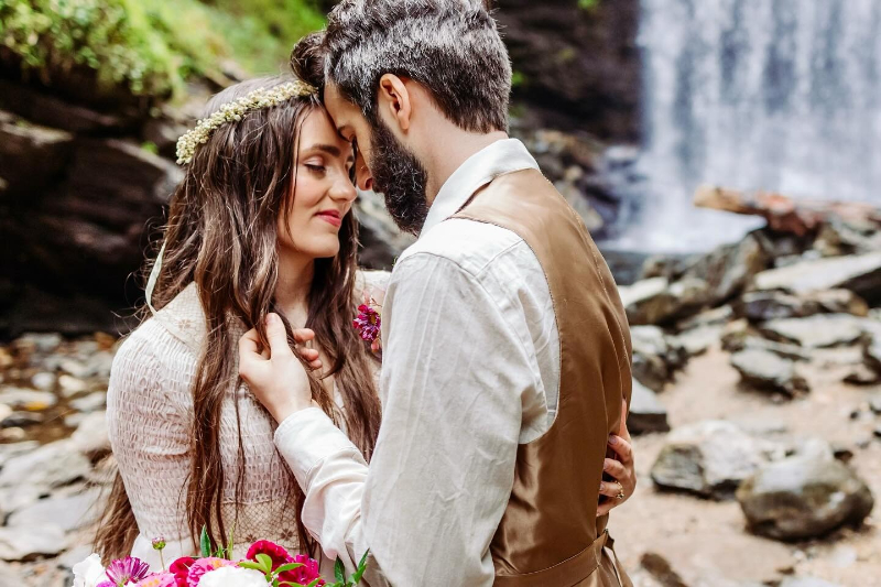Couple sharing a quiet moment in front of a waterfall, surrounded by rocks and lush forest greenery.