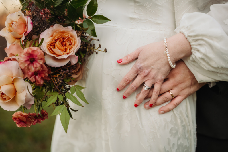 Close-up of couple’s hands resting on a wedding dress, with a bouquet of peach and pink roses.