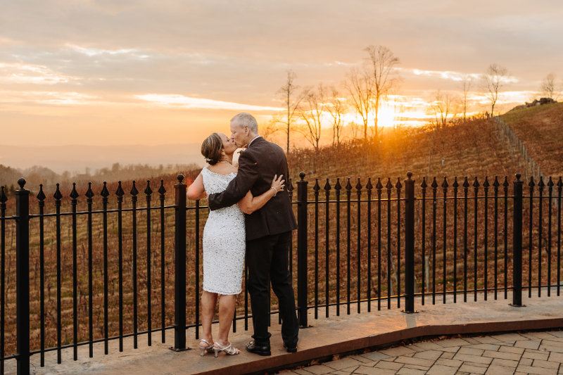 Couple shares a kiss at sunset on a scenic terrace overlooking rolling hills and bare trees.