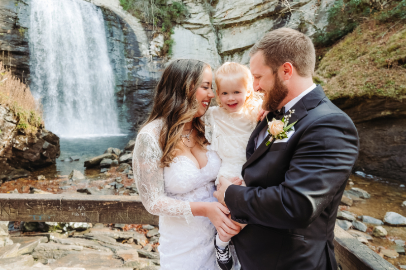 Smiling couple with child in wedding attire by a waterfall, showing what to expect the week of your elopement.