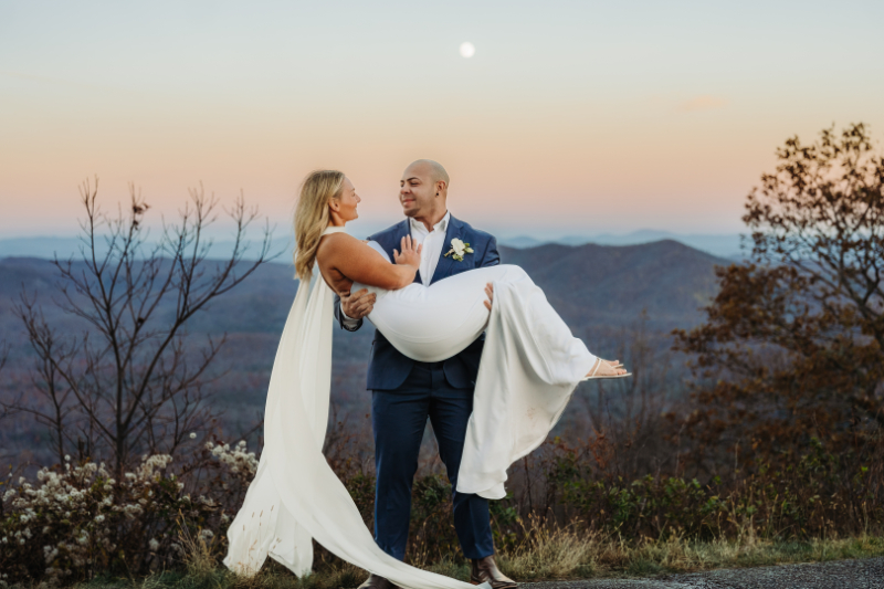 Groom carrying bride in front of Blue Ridge Mountains at sunset during their elopement and honeymoon combo.