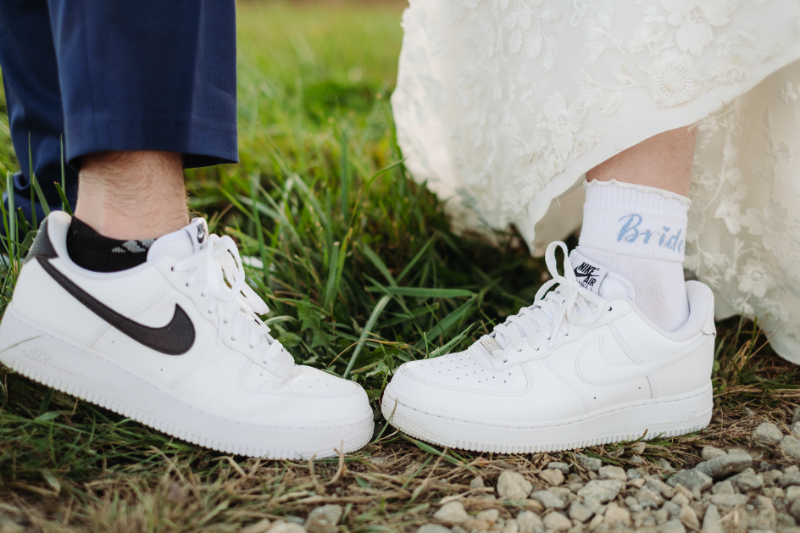 Close-up of bride and groom wearing matching white sneakers, with "Bride" on her socks and grass beneath.