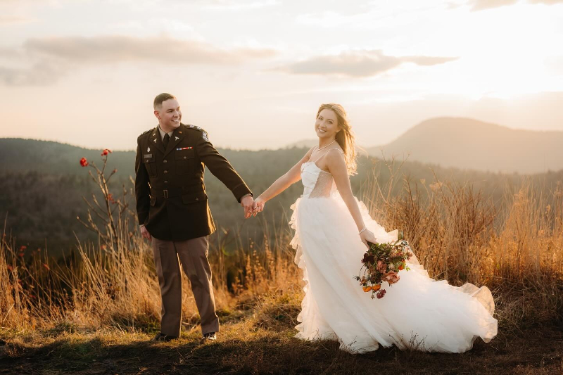 Bride and groom hold hands at sunset on a mountaintop, with golden light and scenic views.
