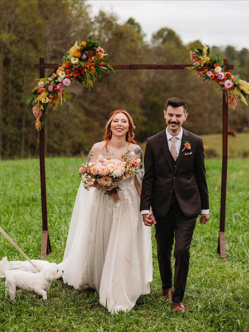 Joyful couple walks hand in hand after outdoor ceremony, surrounded by flowers and lush green meadow.